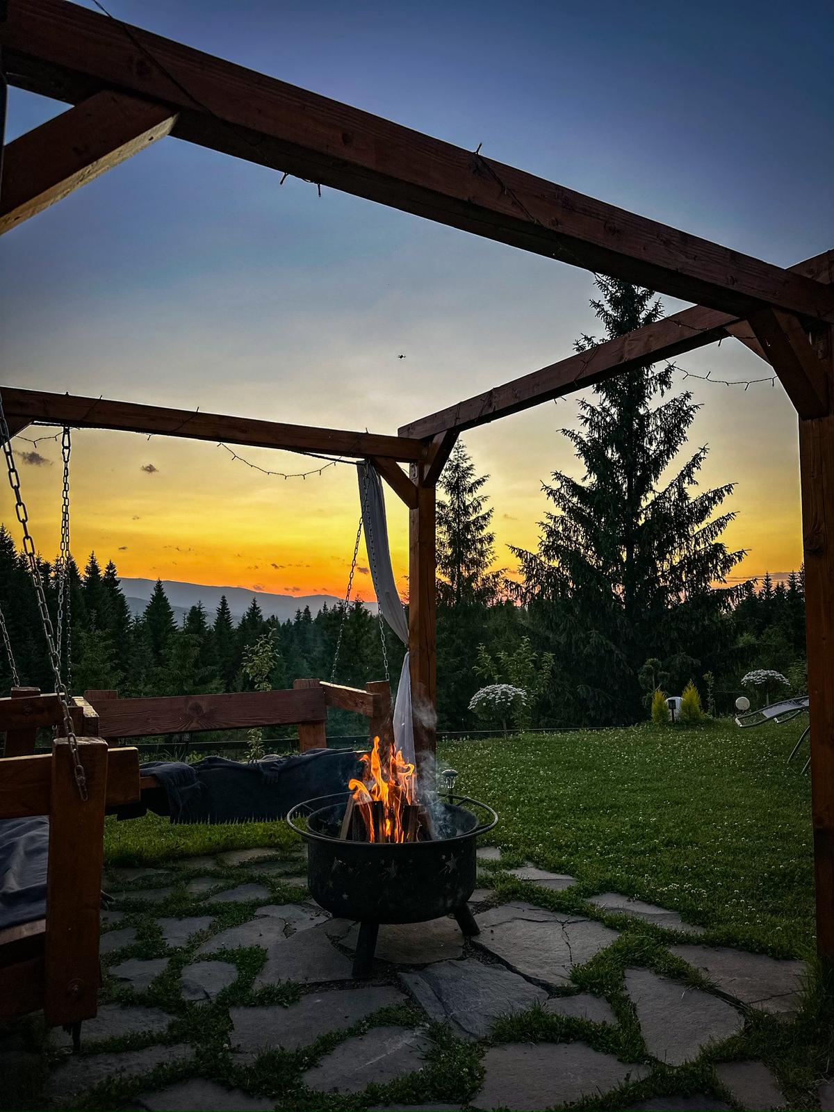 Outdoor fire pit at sunset with swing bench and mountain forest views under wooden pergola